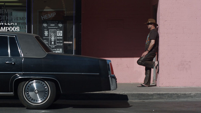 A man wearing a cowboy hat rest agains a pink wall next to a black car