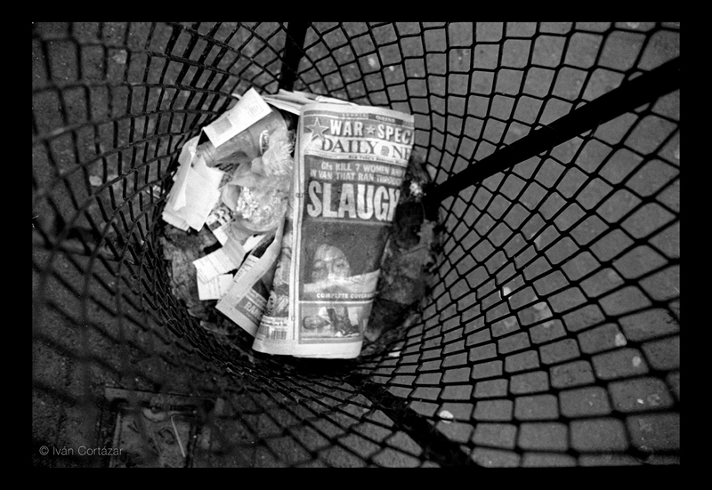 A black and white photo of a newspaper with Iraqi war headlines and imagery inside a wired trash can.
